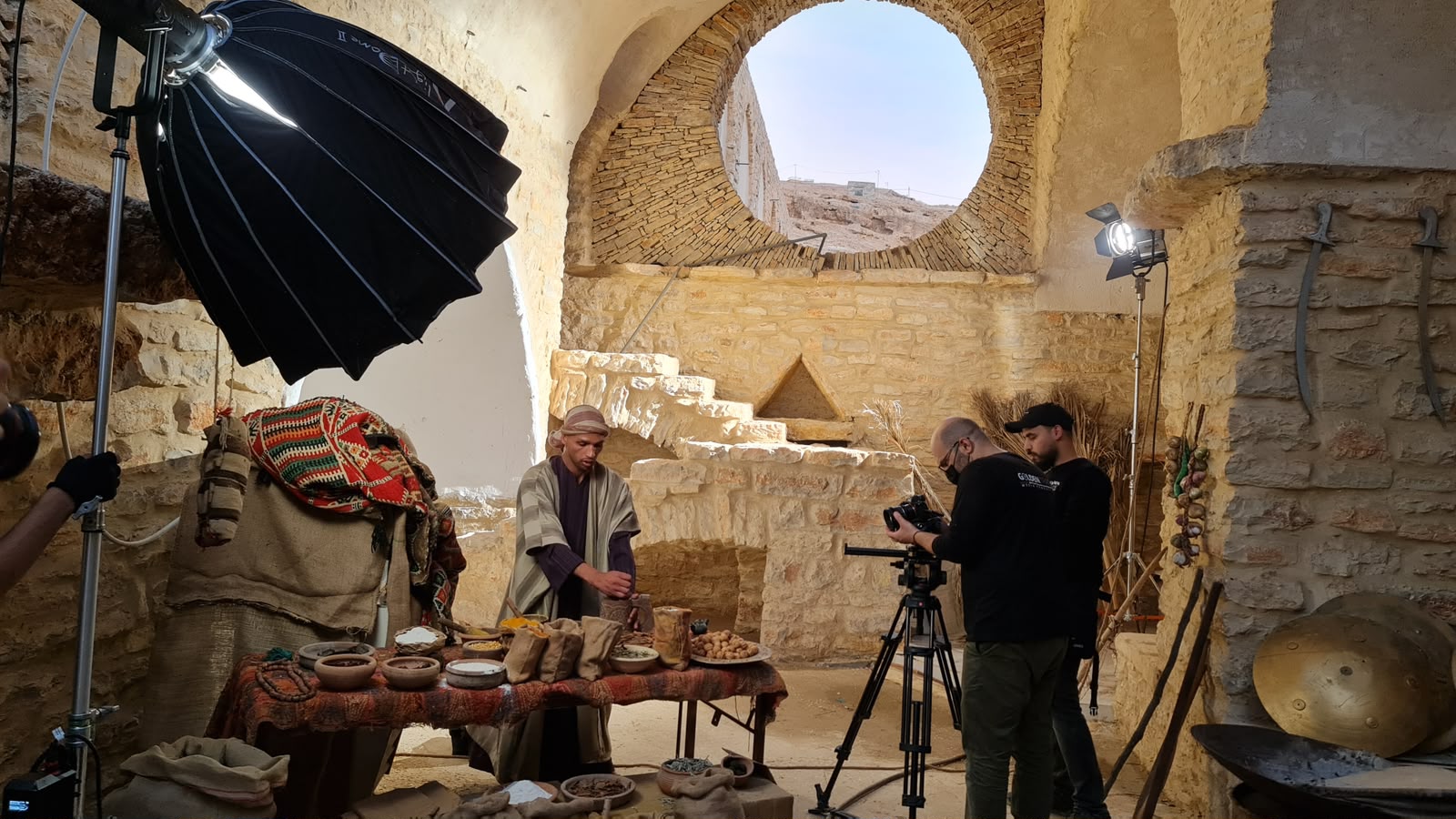 موقع تصوير بقبو حجري مفروش بالتوابل والأكياس، ممثل بالزيّ عند الطاولة والطاقم يصوّر